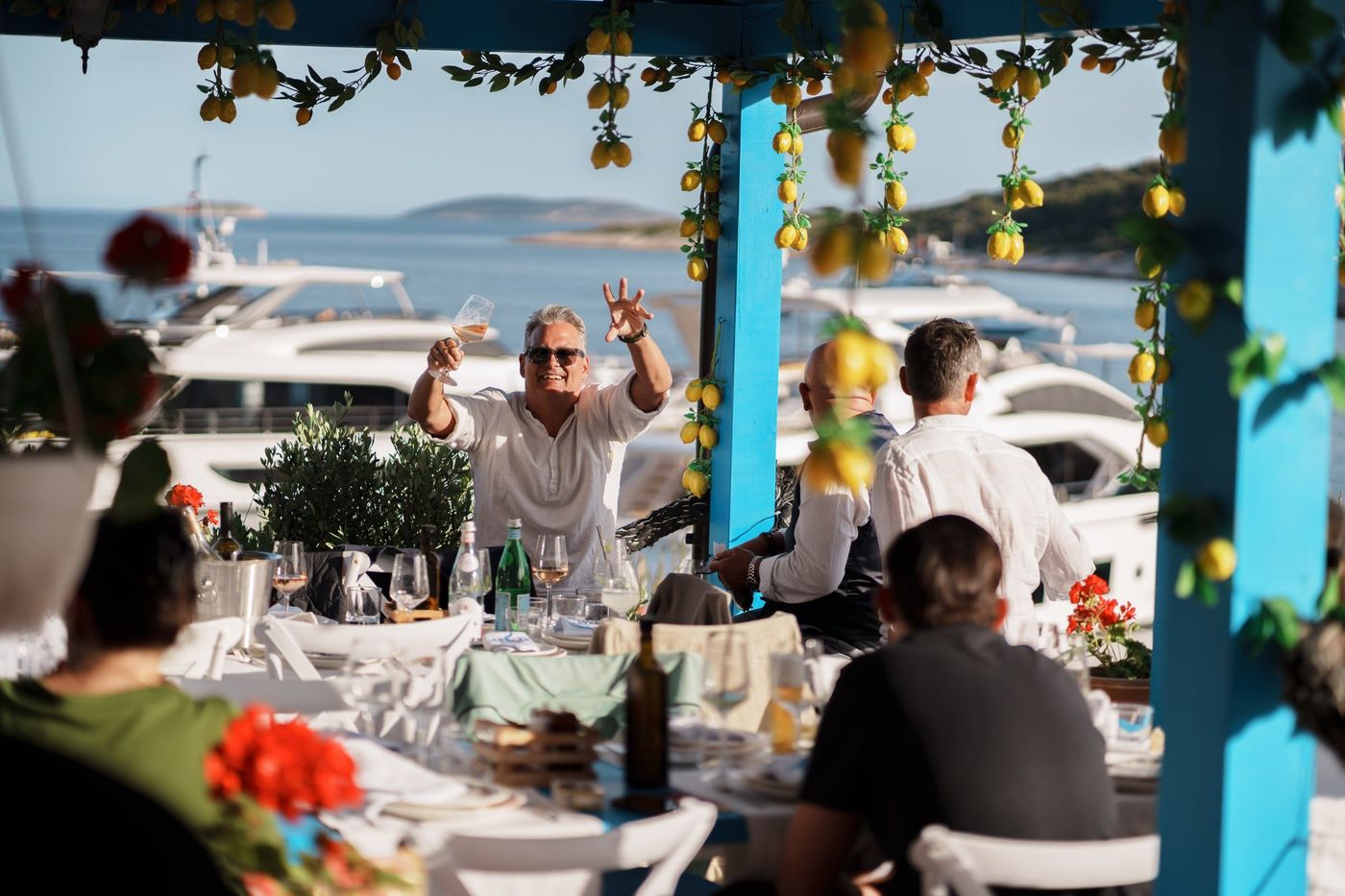 A guest raising a glass at a sunlit table on the terrace, yachts moored in the bay behind.